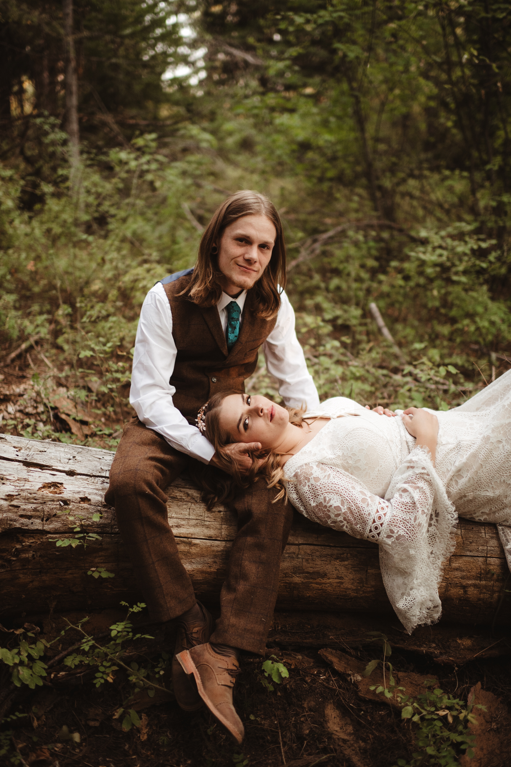 Romantic bride and groom in a serene forest setting, with the bride resting her head on the groom’s lap while lying on a fallen log.